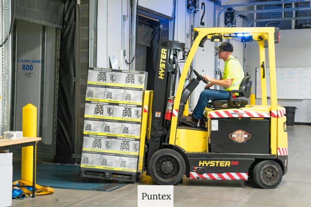 A worker drives a Hyster forklift moving Joe IPA boxes in a warehouse.